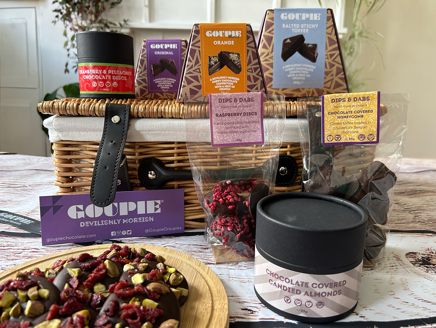 Assorted snacks and treats including a wicker basket, chocolate-covered almonds, and Goopie brand products on a wooden surface.