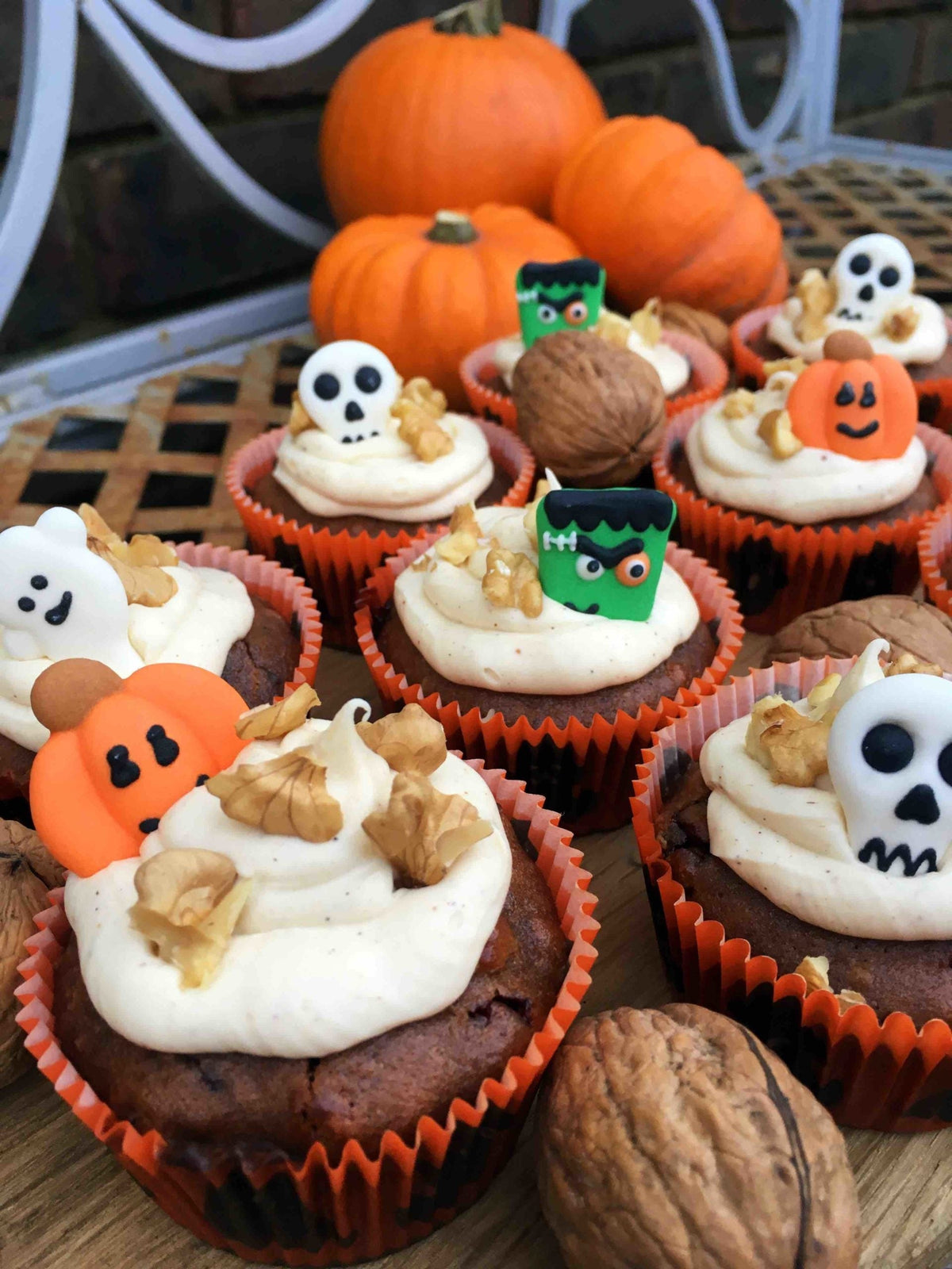 a display of pumpkin spice muffins, each of which is topped with cream cheese frosting and halloween-themed sugar decors like skulls, pumpkins, and zombie heads