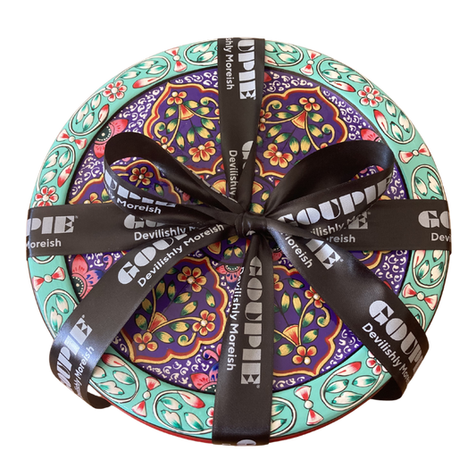 A round decorative gift tin with a colourful floral and paisley pattern, wrapped with a black ribbon printed with the brand name 'GOUPIE' and tagline 'devilishly moreish'