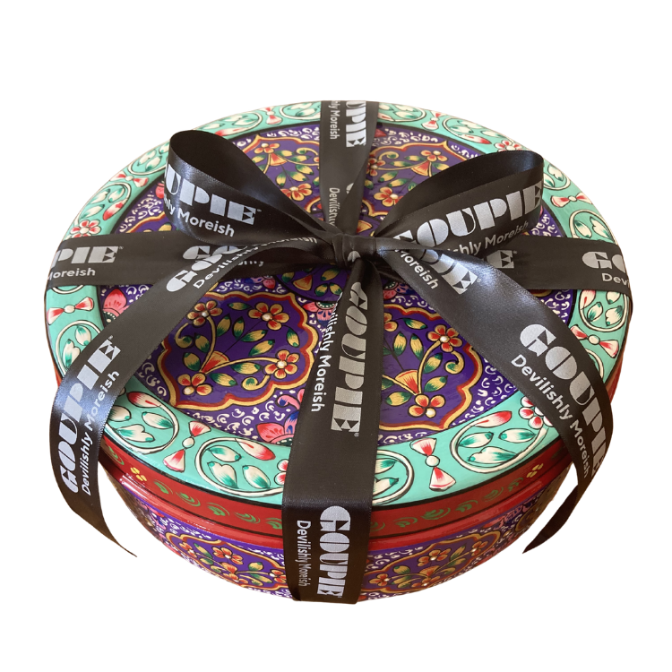 Sideview of a round decorative gift tin with a colourful floral and paisley pattern, wrapped with a black ribbon printed with the brand name 'GOUPIE' and tagline 'devilishly moreish'