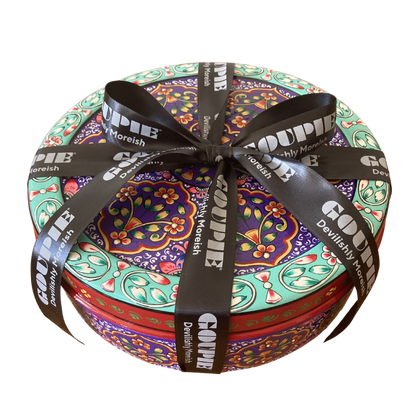 Sideview of a round decorative gift tin with a colourful floral and paisley pattern, wrapped with a black ribbon printed with the brand name 'GOUPIE' and tagline 'devilishly moreish'